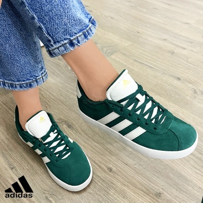 Ténis Adidas verdes em camurça com riscas brancas e sola branca em chão de madeira