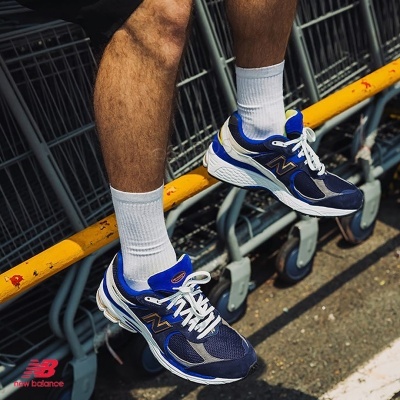 Ténis New Balance em azul escuro, cinzento e branco, com meias brancas próximos de carrinhos de supermercado.