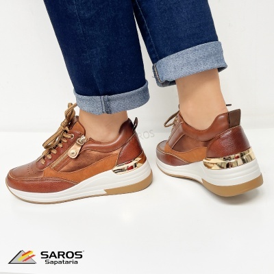 Ténis castanho com detalhes dourados e sola branca, usados com calças de ganga azul escuro.