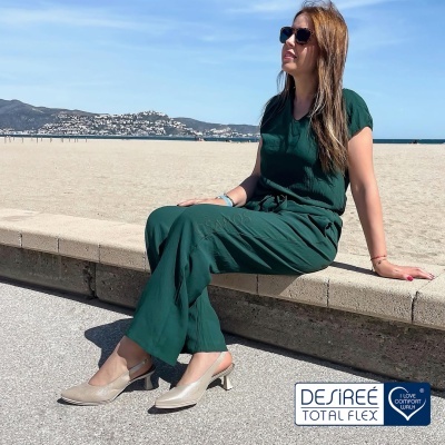 Mulher com macacão verde escuro e sapatos bege sentada numa mureta à beira da praia com logótipo DESIRÉE TOTAL FLEX.