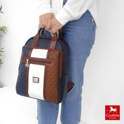 Mochila quadrada azul escuro com detalhes castanhos e faixa branca vertical, sendo segurada por uma pessoa.