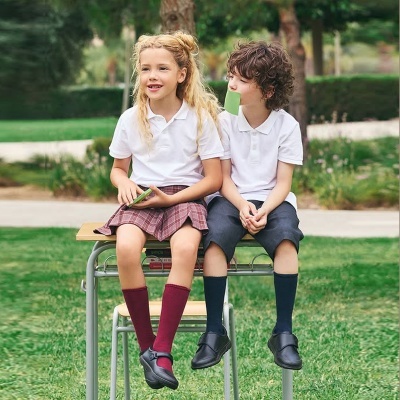 Duas crianças sentadas num banco ao ar livre com uniforme escolar branco e peças de roupa complementares coloridas.