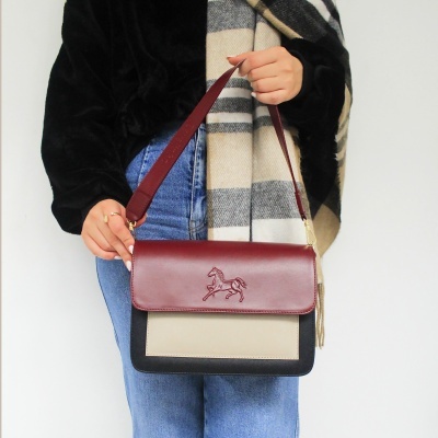 Mala de ombro tricolor com relevo de cavalo, sendo segurada por uma pessoa com casaco preto e jeans