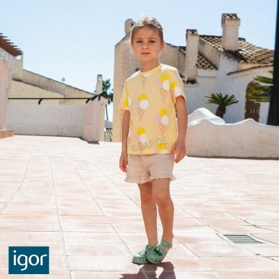 Menina veste t-shirt amarela com gelados, calções beges e sapatos verdes numa azoteia com chão de azulejo