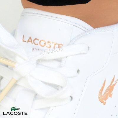Ténis brancos Lacoste com logótipo dourado e texto na língua