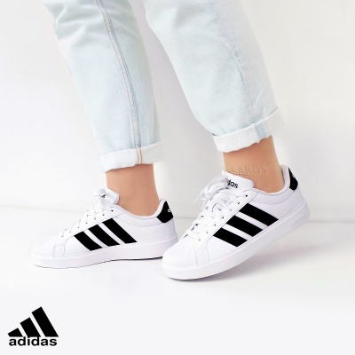 Ténis Adidas brancos com riscas pretas e sola branca usados com calças de ganga claras