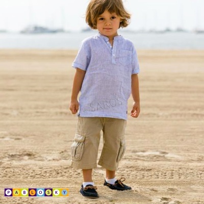 Criança na praia com camisa azul clara de manga curta, calças cargo bege e sapatos pretos e brancos