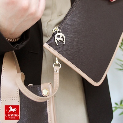 Bolsa de mão bicolor castanho e bege com ferradura dourada e pendente, mão segurando e roupa formal