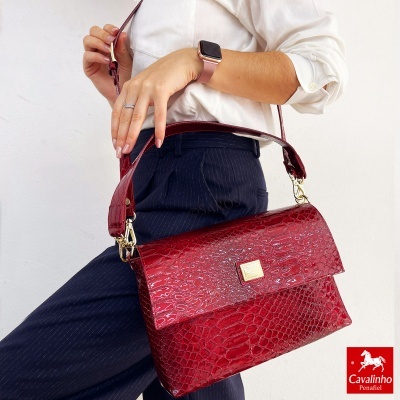 Mala vermelha de couro padrão crocodilo com fecho e plaqueta dourada, usada por uma pessoa com roupa formal.