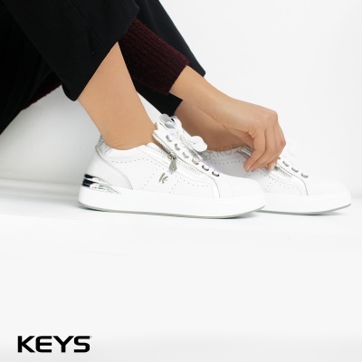 Ténis brancos em pele com detalhes prateados junto a pernas com calças pretas e meias castanhas