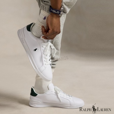 Ténis brancos Polo Ralph Lauren com pormenores verdes e logo preto, usados com meias brancas e calças cinza.
