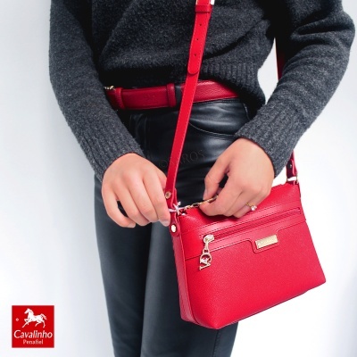 Mala vermelha pequena em pele com fecho zip e pendente dourado, usada por pessoa com roupa preta e cinzenta