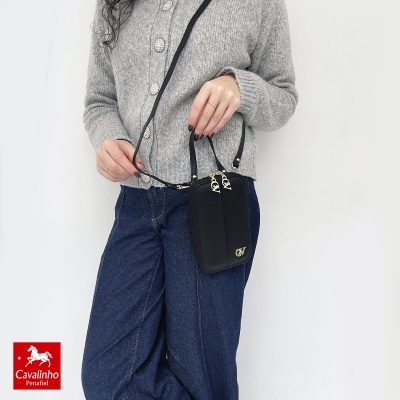 Bolsa pequena preta com alça fina e fecho dourado na mão de uma mulher em moletom cinza e jeans azul