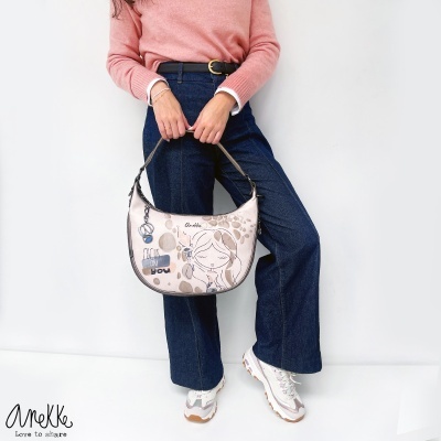 Mala bege ilustrada da marca Anekke com padrão de manchas castanhas e chaveiro metálico, segurada por mulher com camisola rosa e calças jeans.