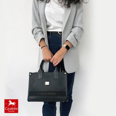 Mala preta de mão em pele com alças e pormenores, com gravação CAVALINHO segurada por pessoa com casaco cinza