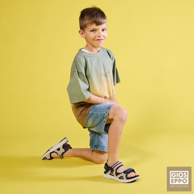 Rapaz infantil de camisa degradê verde-bege, calções de ganga azuis e sandálias desportivas, fundo amarelo