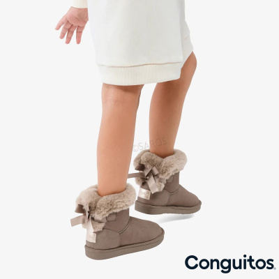 Botas infantis beges com forro felpudo e fita castanha usadas por criança com vestido creme