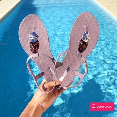Sandálias lilás com decoração floral seguradas em frente a uma piscina azul com etiqueta Ipanema
