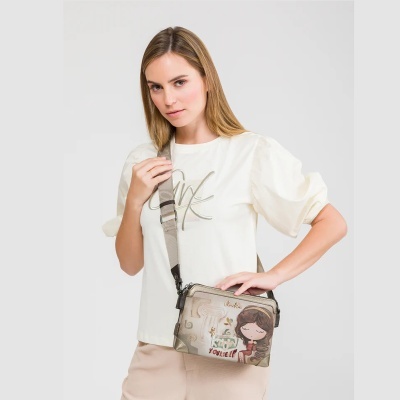 Mala tiracolo bege com alça cinzenta e ilustração frontal, usada por mulher com camisa branca.