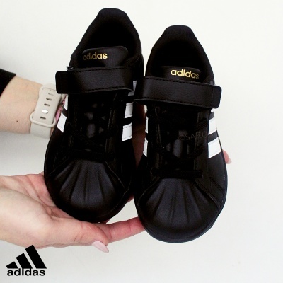 Par de sapatilhas Adidas pretas com riscas brancas e fecho de velcro numa mão