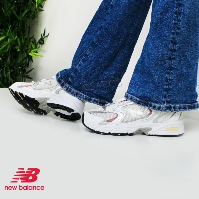 Ténis New Balance brancos com detalhes cinzentos e vermelhos usados com calças de ganga azuis.