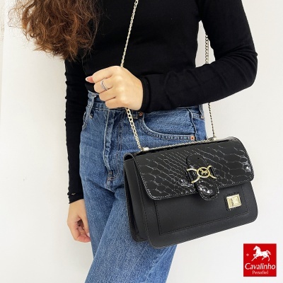 Mala preta com padrão crocodilo e detalhes dourados, com alça de corrente, usada por mulher