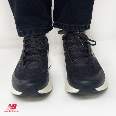 Sapatilhas New Balance pretas com detalhes bege e branca na sola usadas por pessoa com calças pretas