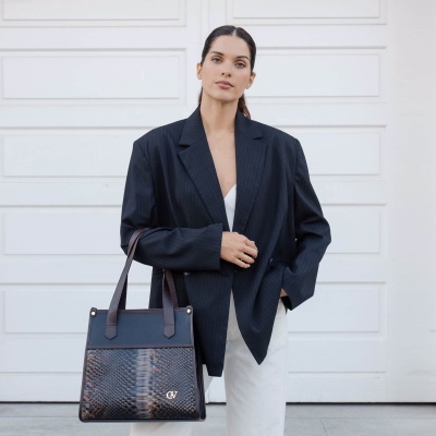 Mulher com blazer preto às riscas segurando bolsa padrão pele de cobra castanha e preta com letras douradas CV