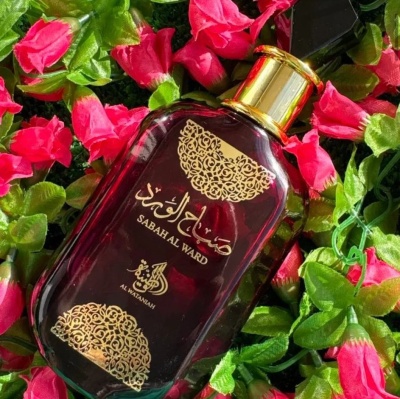 Frasco de perfume vermelho escuro com decoração dourada entre flores artificiais vermelhas.