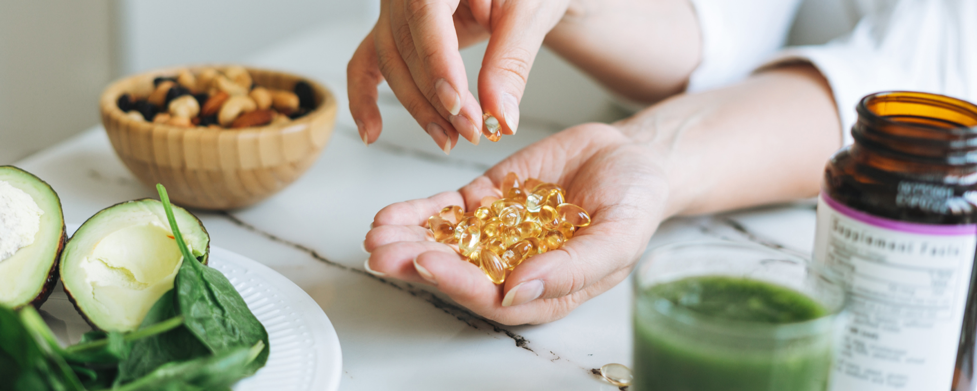 Mão segurando cápsulas gelatinosas amarelas próximas a frasco de suplemento e ingredientes naturais sobre mesa