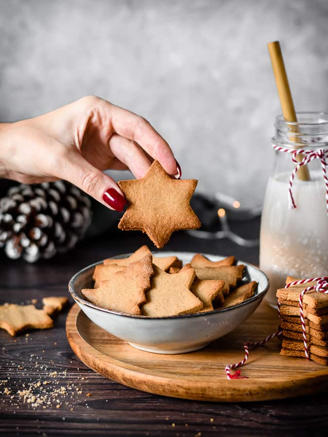 Mão segurando biscoito estrela sobre tigela com biscoitos star e árvore ao lado leite em frasco com canudo e pilha de biscoitos num tabuleiro de madeira.