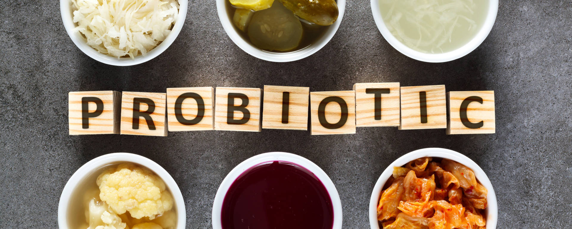 Blocos de madeira com letras formando a palavra PROBIOTIC rodeados por taças brancas com alimentos fermentados.