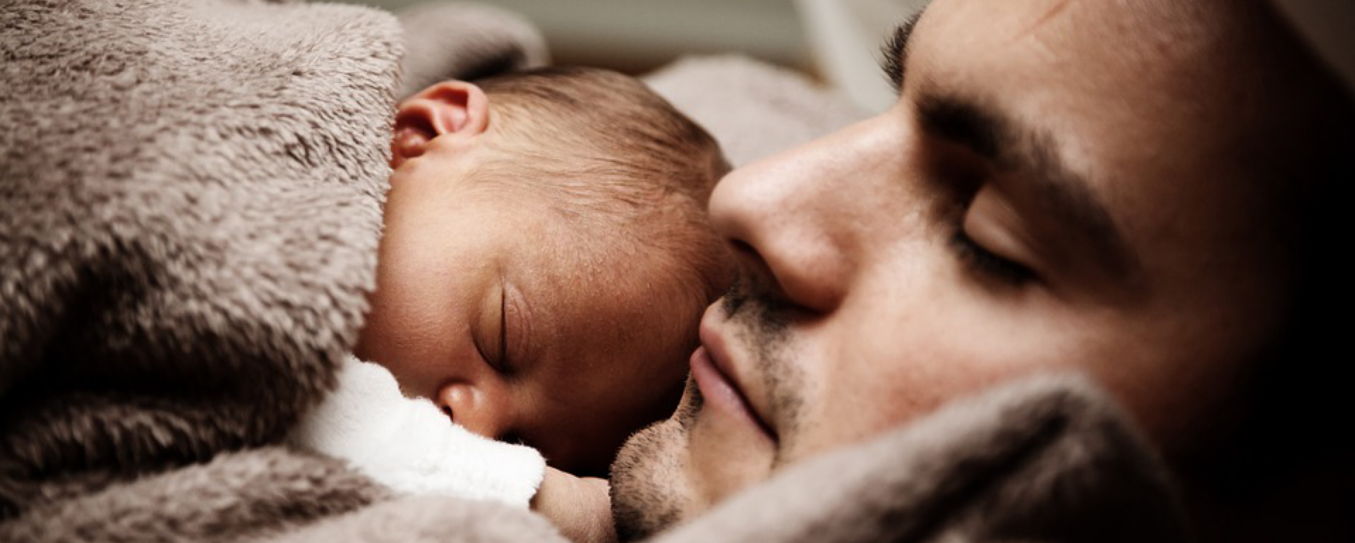 Bebé adormecido com manta castanha ao lado do rosto de um adulto masculino de olhos fechados