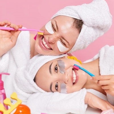 Duas jovens com roupões e toalhas na cabeça usando máscaras faciais e escovas de dentes coloridas num fundo rosa com produtos de cuidado pessoal à frente.