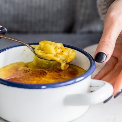 Creme amarelo queimado numa taça branca com bordo azul escuro, com colher e mãos de unhas pretas.