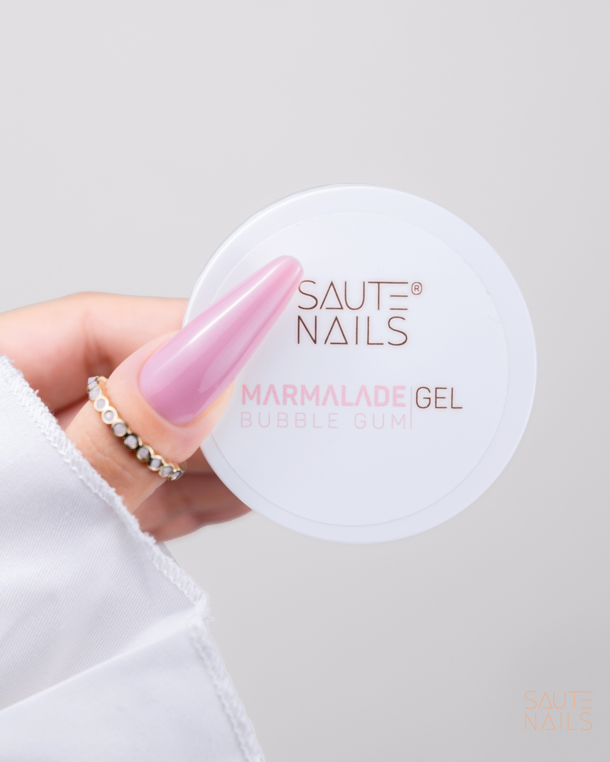 Gel Construtor Marmalade Bubble Gum (Rosa Frio) 30g Frasco branco de gel para unhas SAUTE NAILS Marmalade Gel Bubble Gum segurado por mão com unhas rosas e anéis.