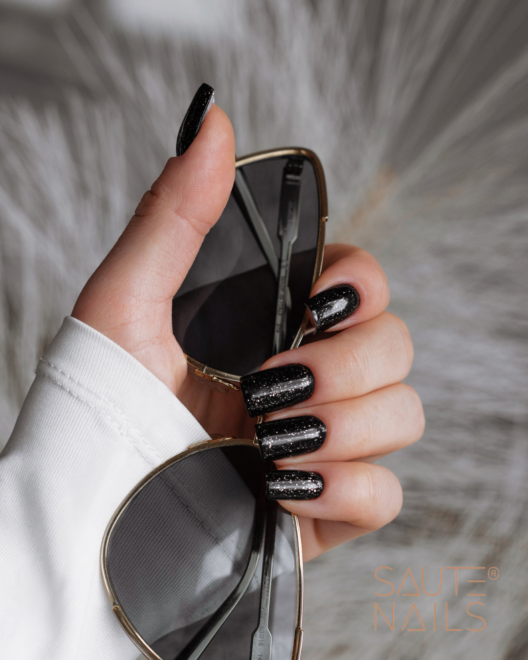 Glimmer Top No Wipe (Partículas de Ouro Brilhantes) 8ml Mão com unhas pretas brilhantes segurando óculos de sol dourados com lentes escuras