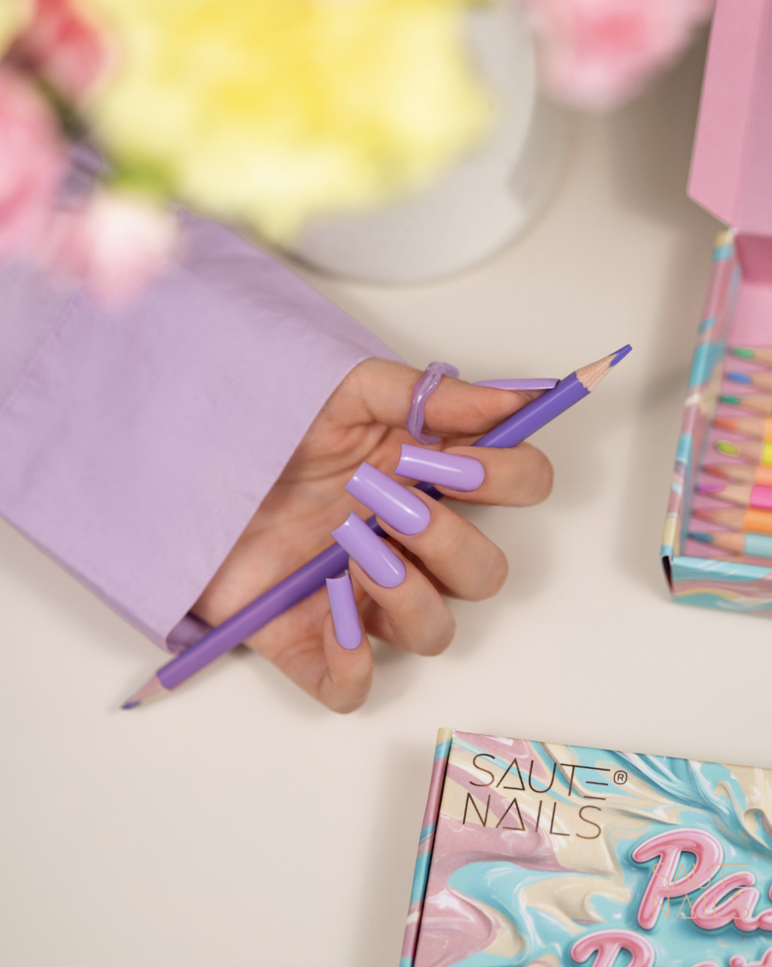 Verniz Gel S294 - Misty Blue (Azul Pastel) Mão com unhas lilás segurando lápis lilás junto a embalagem colorida com texto SALUTE NAILS