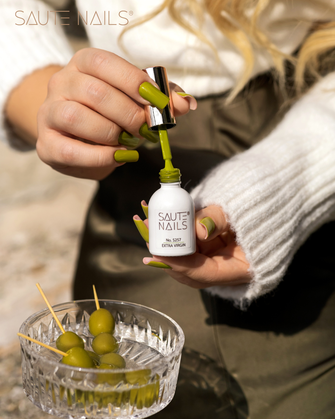Verniz Gel S257 Extra Virgin (Verde Oliva) Verniz para unhas verde SAUTE NAILS com mão feminina e azeitonas verdes em tigela de vidro