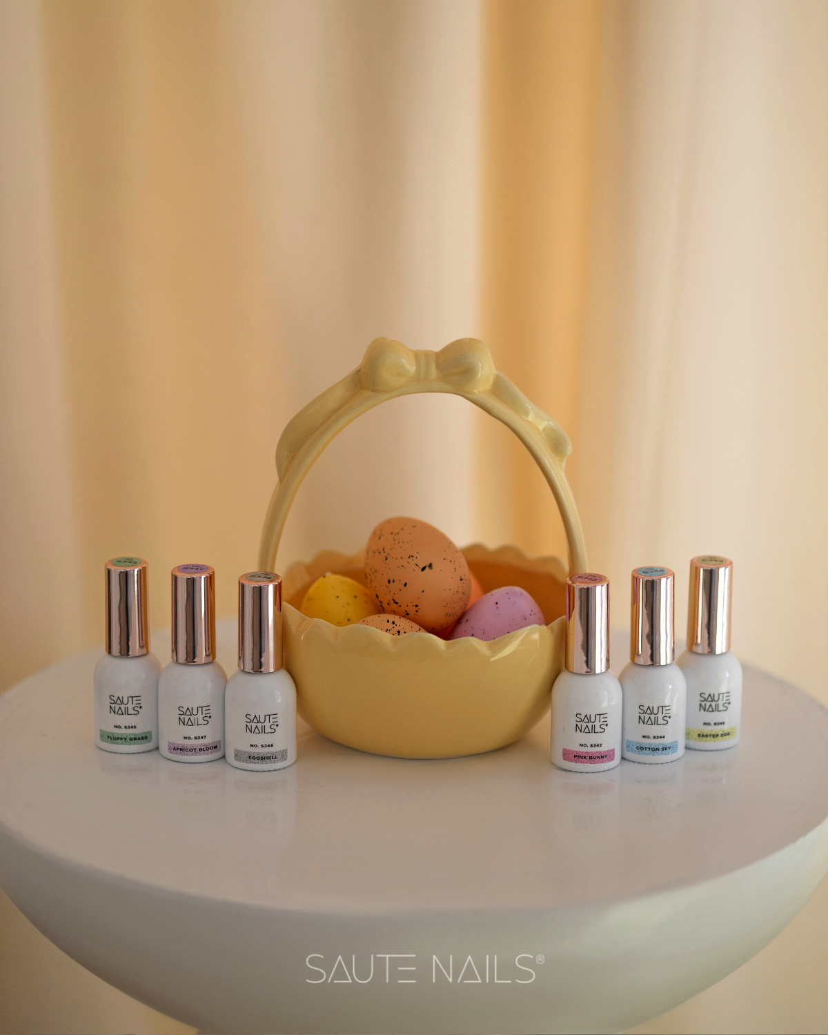 Coleção de Verniz Gel Pastel Eggshell Vernizes SAUTE NAILS em frascos brancos com tampas douradas junto a um cesto amarelo com ovos coloridos