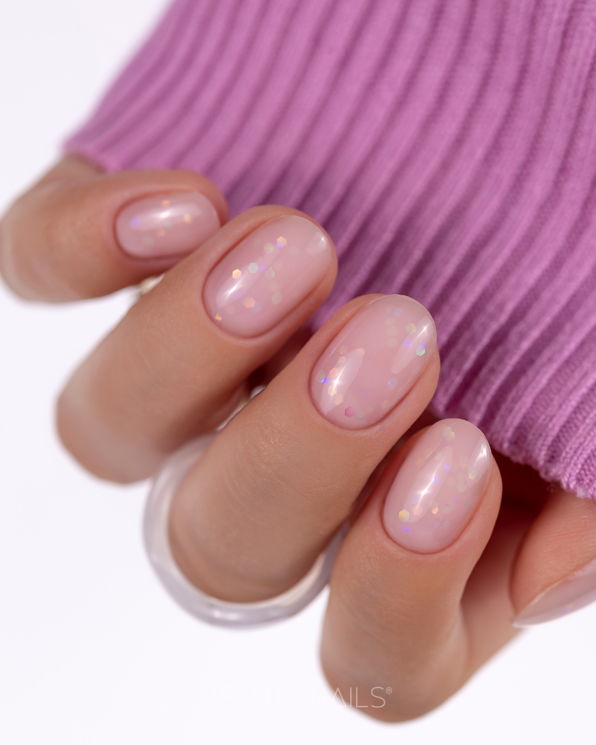 Luna Base Aura (Rosa Leitoso com Partículas Holográficas) Mão com unhas pintadas de rosa claro com glitter sobre tecido roxo de malha