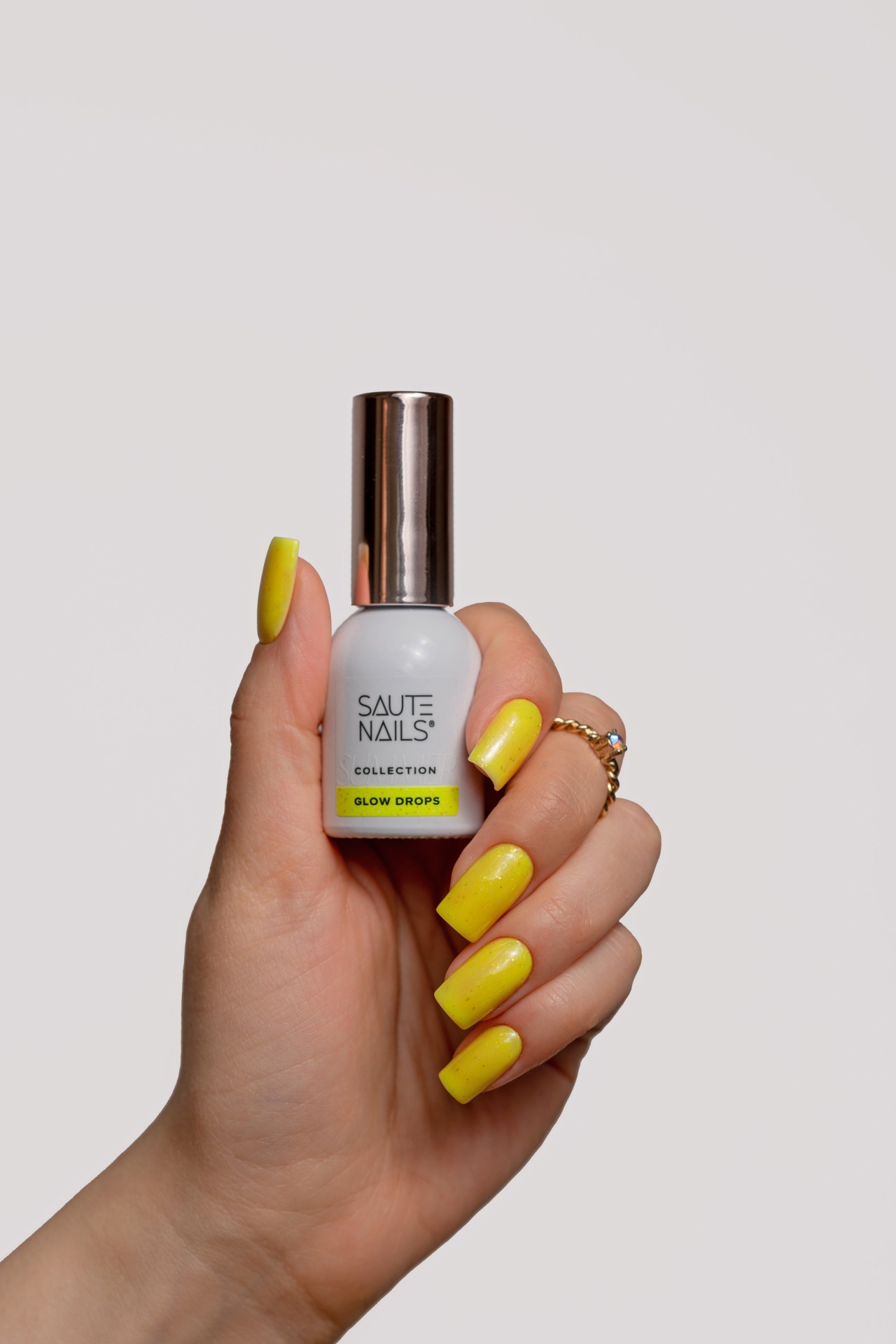 Verniz Gel Glow Drops (Amarelo Intenso com Partículas Douradas) Unhas amarelas segurando verniz SAUTE NAILS glow drops