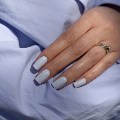 Unhas compridas quadradas brancas com pequenas manchas pretas em mão com anel dourado sobre tecido azul claro