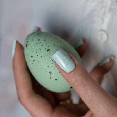 Mãos com unhas azul claro com pontos pretos seguram ovo verde com manchas pretas junto a camisa branca.