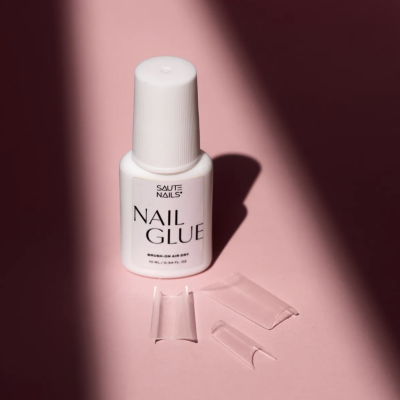 Cola para unhas branca com três unhas postiças transparentes sobre fundo rosa.