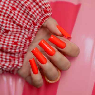 Unhas artificiais vermelhas alaranjadas em mão com blusa xadrez sobre fundo rosa