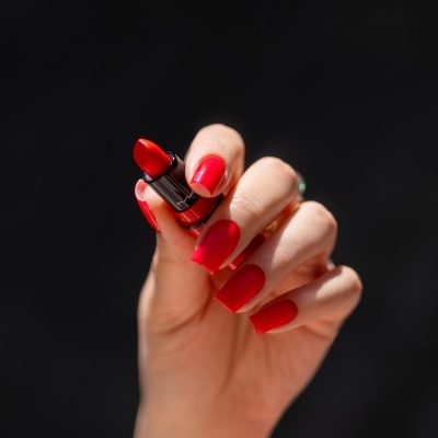Mão feminina segurando batom vermelho com unhas vermelhas, fundo preto