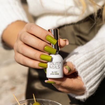 Mãos segurando verniz Saute Nails cor verde oliva com azeitonas ao fundo