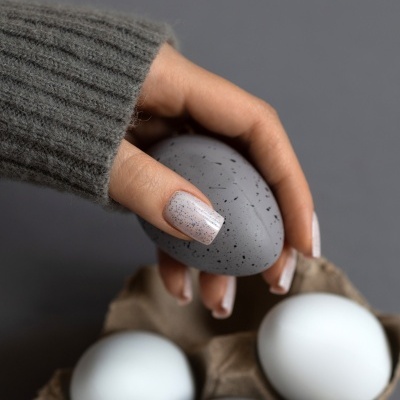 Mão segurando ovo cinzento manchado com unhas pintadas nude manchadas sobre embalagem de ovos brancos e cinzentos.