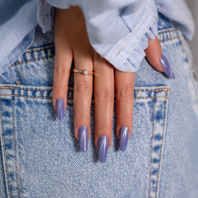 Mão feminina com unhas lilás cintilantes e anel, apoiada em calças de ganga azul.